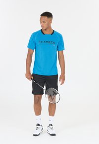 Blå idrottströja med svarta detaljer och logotyp, kombinerad med svarta shorts. Personen håller ett badmintonracket och har på sig vita strumpor och skor.