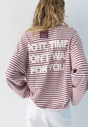 Personne aux longs cheveux bouclés portant un t-shirt à manches longues rayé blanc et violet avec le texte "FAIS-LE, LE TEMPS N'ATTEND PAS" au dos.