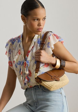 Kobieta w kwiecistej bluzce i jasnych dżinsach trzyma plecioną słomianą torebkę na ramię z brązową skórzaną klapką i paskiem, ozdobioną złotymi i żółwiowymi bransoletkami.