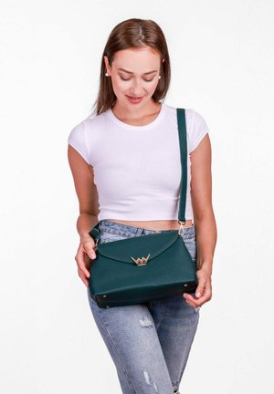 Zelená kabelka z umělé kůže na rameno se zlatým akcentem na klipsu, obdélníkového tvaru a nastavitelným popruhem, kterou drží osoba v bílém topu.