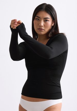 Woman with straight dark hair wears a fitted black long-sleeve top and white underwear, adjusting her right sleeve with her left hand.