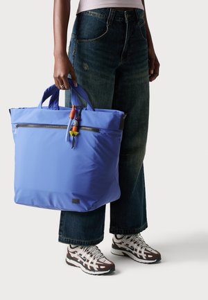 Stor blå tote taske lavet af nylon, med to håndtag, en frontlomme med lynlås og farverige dekorative charms. Glat tekstur.
