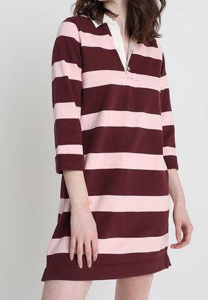 Frau trägt ein knielanges Polo-Kleid mit langen Ärmeln, das breite horizontale Streifen in Burgunderrot und Pink auf einem einfarbigen Hintergrund zeigt.