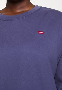 Primo piano di una persona che indossa una maglietta blu a girocollo con un piccolo logo rosso di Levi's sul lato sinistro del petto.