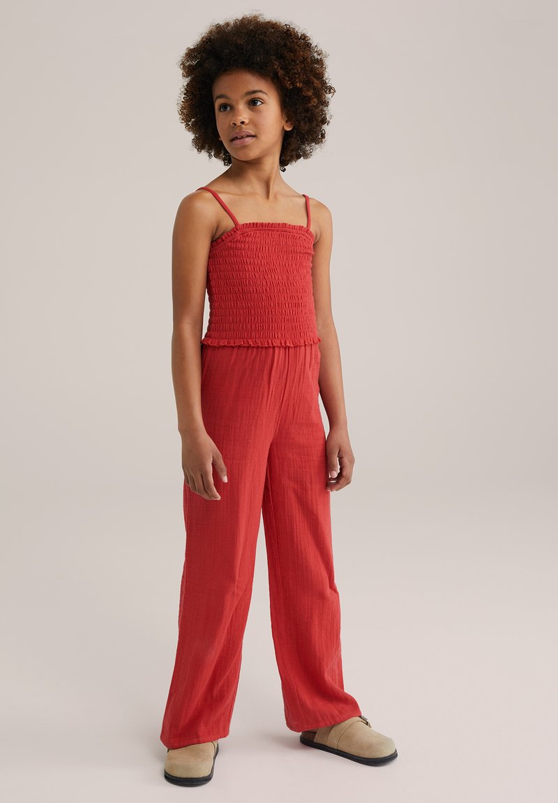 Red woven jumpsuit with smocked bodice and wide-leg pants, featuring thin straps and a textured fabric. Model is wearing beige slip-on shoes.
