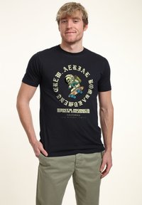 Černé bavlněné tričko s barevným grafikou lebky v astronautském obleku, jak skateboarduje, s textovými prvky v žluté a bílé barvě.