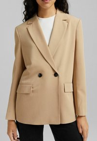 Beżowa dwurzędowa marynarka z klapami, dwoma czarnymi guzikami i bocznymi kieszeniami. Gładki materiał o dopasowanym kroju.