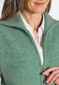 Grüner gerippter Strickwesten mit hohem Kragen und Reißverschluss, getragen über einem weißen Hemd. Der Stoff hat eine weiche Textur mit bläulichen Tönen.