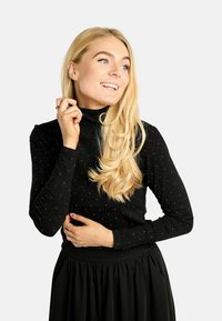 Czarna bluzka z długim rękawem i wysokim kołnierzem, zdobiona błyszczącym wzorem. Tkanina ma gładką fakturę, uzupełnioną przez zwiewną czarną spódnicę.