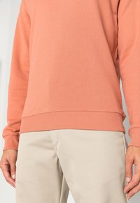Katoenen sweatshirt in koraal met ribgebreide boorden en zoom, gecombineerd met beige textuurpantalons. Focus op stof- en pasvormdetails.