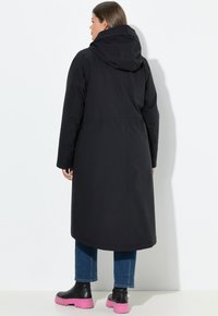 Dlouhý černý voděodolný kabát s kapucí, jednoduše navržený a volného střihu, v kombinaci s modrými džínami a černo-růžovými platformovými botami.