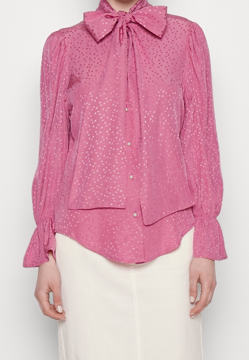 Rosa blus med en fluga vid kragen, långa puffärmar och ett subtilt geometriskt mönster. Knäppt med knappar, tillverkad av lättviktsmaterial.