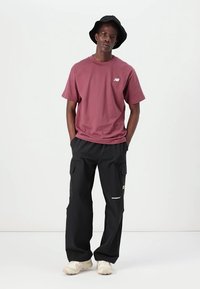 Bordeauxfarbene Baumwoll-T-Shirt mit kleinem Logo, kombiniert mit schwarzen Cargo-Hosen mit mehreren Taschen und beigen Sneakers. Schwarzer Bucket Hat.