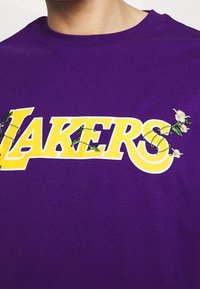Fialové bavlněné tričko s nápisem "LAKERS" v žluté barvě. Text má květinovou výšivku a zelené akcenty, které doplňují výrazný design.
