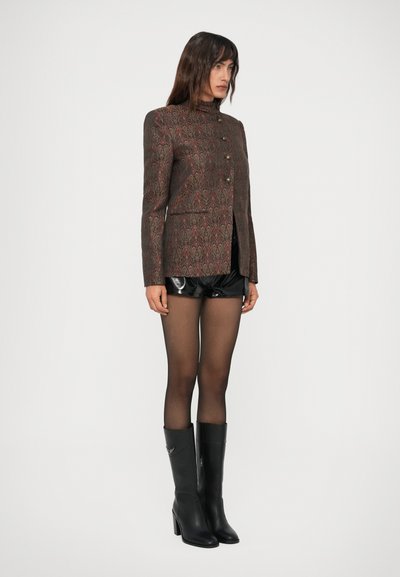 Femeie îmbrăcată cu un sacou maro cu model, pantaloni scurți negri lucioși, ciorapi negri transparenți și cizme negre până la genunchi, stând în fața unui fundal alb.