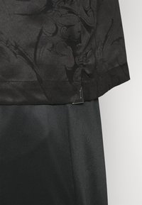 Musta vaate, jossa on sileä rakenne ja sävyttävä abstrakti kuvio, jota korostaa metallinen kolmion yksityiskohta helman kohdalla.