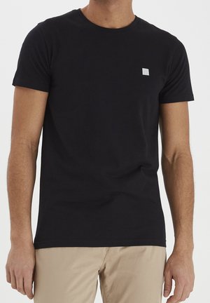 Man draagt een effen zwart T-shirt met korte mouwen met een klein wit vierkant logo op de borst en beige broek, staand tegen een witte achtergrond.