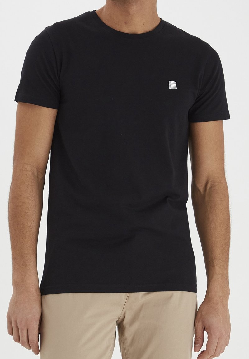 Homme portant un t-shirt noir uni à manches courtes avec un petit logo carré blanc sur la poitrine et un pantalon beige, debout devant un fond blanc.