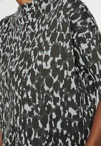 Pletený top s vysokým krkem, krátkými rukávy a texturovaným vzorem tmavě zelených a světlých šedých odstínů uspořádaných v abstraktním designu.