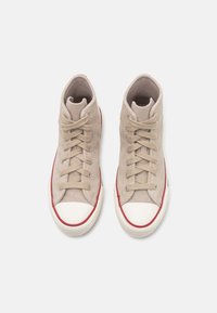 Zapatillas altas beige de ante con puntera blanca, que presentan cordones redondos, acentos rojos y paneles laterales texturizados.