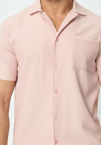 Chemise rose pâle à manches courtes avec une texture en relief verticale, un devant à boutons et une poche. Tissu doux avec de petits boutons ronds.