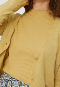 Yellow ribbed turtleneck with textured yellow cardigan featuring round buttons. Background shows a patterned skirt with floral design.