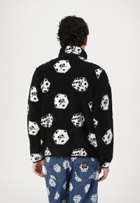Black fleece jacket featuring white and black dice patterns, high collar, and a soft texture. Paired with light blue jeans with similar designs.