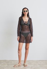 Svart mesh cover-up med lange ermer, V-hals og knytefront, lagt over en mørk bikini. Modellen har på seg grønne solbriller og beige sandaler.