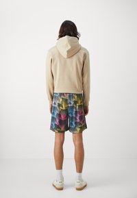 Felpa beige con cappuccio e coulisse, abbinata a pantaloni shorts multicolori con motivi che presentano sfumature di rosa, blu e giallo e un design testurizzato. Sneakers bianche.