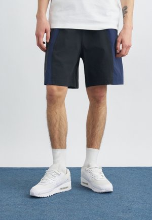 Homme portant un short bicolore noir et bleu marine, des chaussures de sport blanches et des chaussettes blanches montantes, debout sur un tapis bleu.