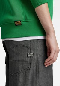 Sudadera verde con puños acanalados, combinada con jeans de mezclilla gris que presentan una etiqueta de G-Star y costuras curvadas en los bolsillos traseros.