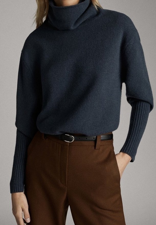 Femme portant un pull à col roulé bleu foncé avec des manches côtelées, rentré dans un pantalon marron taille haute et une ceinture noire.