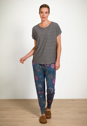 Vrouw die binnenshuis wandelt, gekleed in een gestreepte shirt met korte mouwen, blauwe bloemenbroek en bruine slippers op een houten vloer.