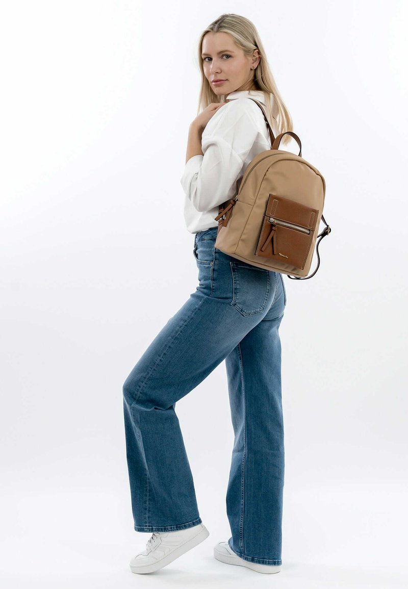 Sac à dos en nylon beige avec une poche avant en cuir marron, fermeture éclair argentée et bretelles réglables, porté par-dessus une blouse blanche et un jean bleu.