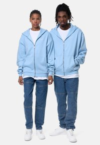 Light blue zip-up hoodies with front pockets, layered over white shirts. Paired with dark blue denim, featuring patchwork sections and white sneakers.