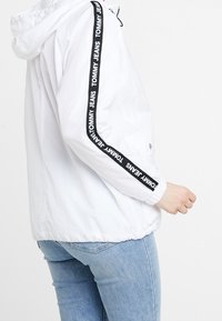 Biała kurtka zapinana na zamek z kapturem, z czarną taśmą z logo na rękawach oraz bocznymi kieszeniami. Lekki, gładki materiał.