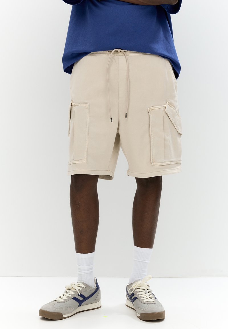 Pantaloni cargo beige con tasche laterali, vita con cordoncino e dettagli di cucitura, abbinati a sneaker grigie e calze corte bianche.