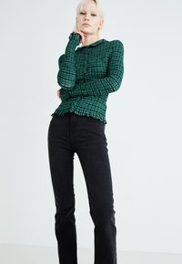 Blusa de manga larga verde con estampado, botones y dobladillo con volantes, combinada con jeans negros. Textura suave, diseño ajustado y detalles de patrón a cuadros.