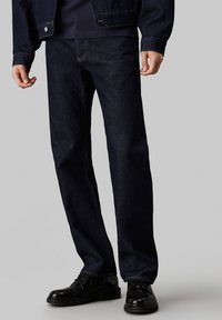 Jean en denim bleu foncé avec une coupe droite, dotés de coutures contrastées et d'une fermeture à bouton, assortis à des chaussures noires.