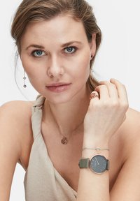 Runde Uhr mit schwarzem Ziffernblatt und grünem Armband, kombiniert mit einer Halskette aus Roségold mit Anhänger-Charm und einem passenden Ring.