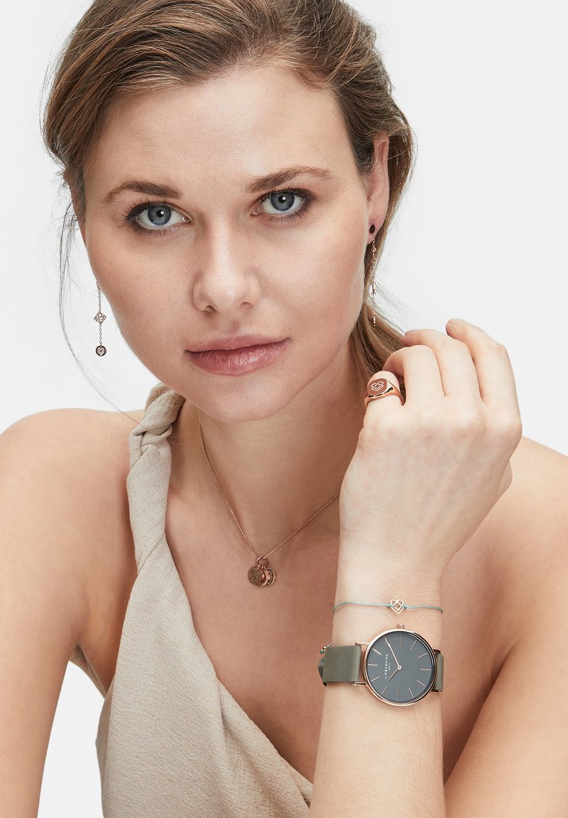 Runde Uhr mit schwarzem Ziffernblatt und grünem Armband, kombiniert mit einer Halskette aus Roségold mit Anhänger-Charm und einem passenden Ring.