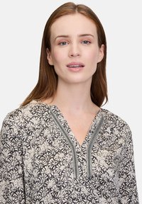 Bluse mit Blumenmuster in Creme und Schwarz mit V-Ausschnitt, ausgestattet mit strukturiertem grauem Besatz entlang der Knopfleiste. Leichtes Material.