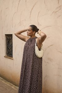 Femme ajustant ses cheveux, portant une robe marron sans manches à pois blancs, avec un petit sac à bandoulière blanc, debout près d'un mur beige texturé.