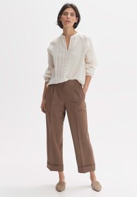 Blusa texturizzata color crema con scollo a V e maniche lunghe, abbinata a pantaloni larghi marroni con risvolto in fondo. Scarpe slip-on di colore marrone chiaro.