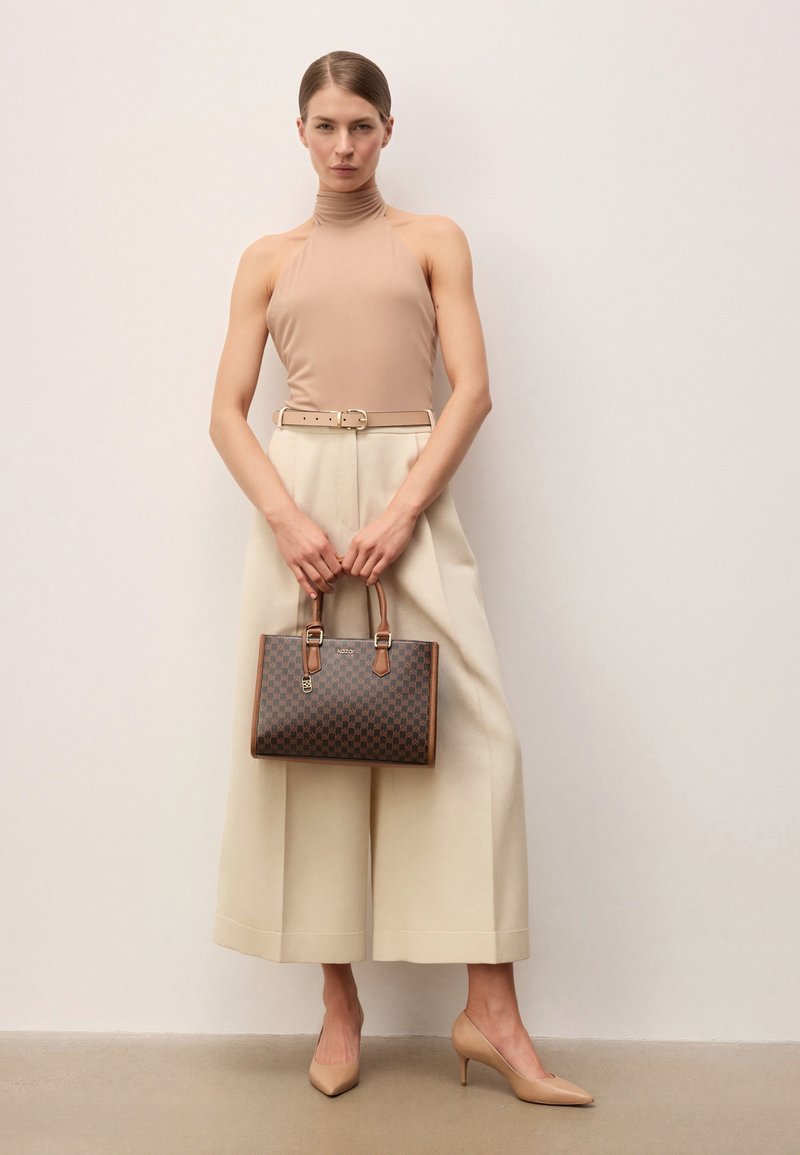 Kobieta w beżowym bezrękawnym golfie i szerokich kremowych spodniach trzyma brązową torebkę w wzory, nosząc beżowe szpilki w szpic.