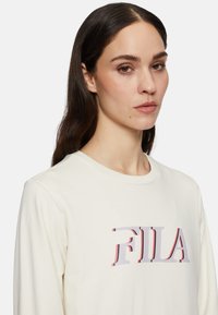 Pitkähihaisista, kerma värisestä puuvillapaitasta. Ominaisuutena näkyvä monivärinen "FILA"-logo rinnassa. Klassinen pyöreä kaula-aukkosuunnittelu.
