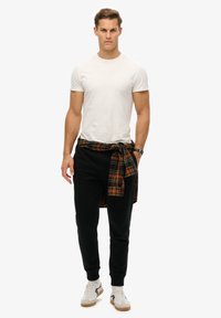 Witte kratärm T-shirt, zwarte getailleerde sweatpants, geruit overhemd in oranje en groen geknoopt om de taille, witte en grijze sneakers.