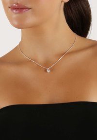 Silverhalsband med en delikat kedja prydd med små runda pärlor och en enda berlock, placerad mot bar hud och en svart topp.