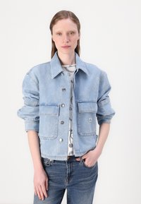 Veste en denim bleu clair avec un col, deux poches avant, fermeture à boutons et manches retroussées. Portée par-dessus une chemise à motif.