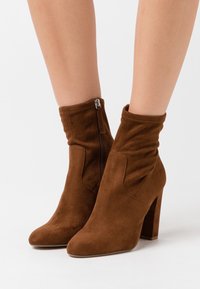 Botas de tobillo de ante marrón con tacones cuadrados y cremalleras laterales, usadas sobre piernas desnudas contra un fondo blanco.